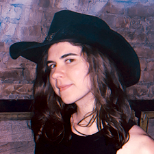 A young, millennial white woman giving a closed-mouth smile and wearing a black cowboy hat. She has brown hair, loosely curled to right below her shoulders.