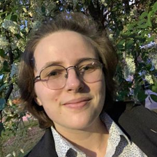 A non-binary person smiling into the camera, their face framed in flowers. They are wearing a white button-down and a black cardigan.